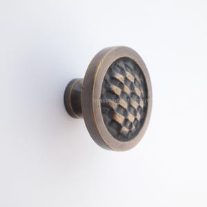 Pode incluir: Um puxador de armário de cor bronze com uma face texturizada com um padrão de losangos. O puxador tem uma forma arredondada com uma face circular e uma base cilíndrica para montagem. O hardware é provavelmente para móveis ou armários.