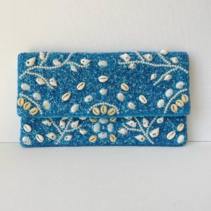 May include: A turquoise beaded clutch with a fold-over flap. The clutch is embellished with white seashells and small white beads, arranged in decorative patterns. The overall design evokes a coastal or nautical theme.