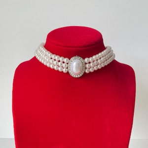 May include: A white pearl choker necklace with three rows of pearls and a large, oval-shaped pearl in the center. The pearl is surrounded by a silver setting with small clear rhinestones.