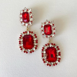 May include: A pair of dazzling red and silver earrings. Each earring features a large, rectangular red gemstone surrounded by smaller clear and red stones. The earrings have a gold-colored connecting chain.