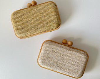 May include: Two rectangular clutch purses. One is a light tan color, and the other is a beige color. Both purses have a woven texture and a wooden frame. Each purse has a wooden clasp with two small, round wooden beads.