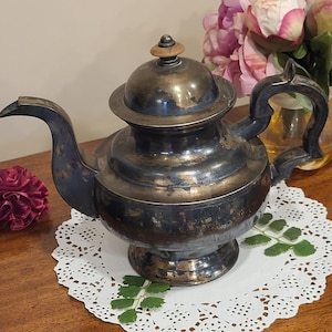 May include: An antique silver-toned teapot with a dark handle and spout. The teapot sits on a white doily with a floral arrangement in the background. The lid has a wooden knob.