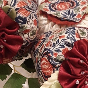May include: Three fabric sachets with a floral pattern in shades of blue, red, and green. Each sachet is tied with a burgundy ribbon and embellished with white beads.