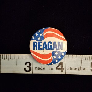 K&ouml;nnte beinhalten: Ein runder Knopf mit patriotischem Design, mit roten und wei&szlig;en Streifen und blauen Sternen. Das Wort "REAGAN" ist wei&szlig; in der Mitte aufgedruckt. Der Knopf liegt neben einem Lineal.