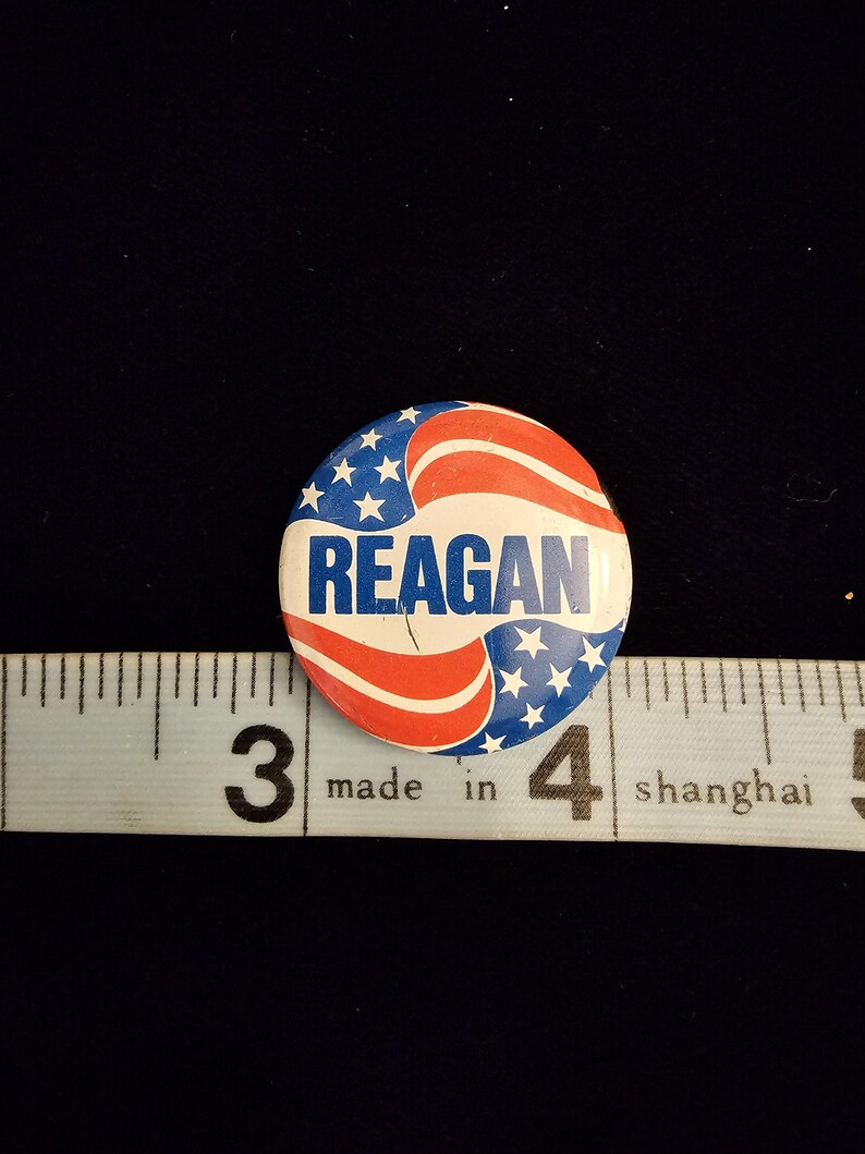 K&ouml;nnte beinhalten: Ein runder Knopf mit patriotischem Design, mit roten und wei&szlig;en Streifen und blauen Sternen. Das Wort "REAGAN" ist wei&szlig; in der Mitte aufgedruckt. Der Knopf liegt neben einem Lineal.