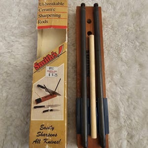 Könnte beinhalten: Ein Messerschärfer-Set der Marke Smith's mit einem rechteckigen Holzsockel, zwei dunkelgrauen Keramikstäben und einem hellen Holzstab. Das Set enthält einen Karton mit der Aufschrift Unbreakable Ceramic Sharpening Rods und Easily Sharpens All Knives.