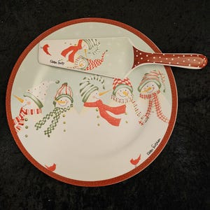 May include: A round, white ceramic plate and matching server with a winter snowman theme. The plate has a red border and features several snowmen with colorful scarves and hats. The server has a red polka-dot handle.
