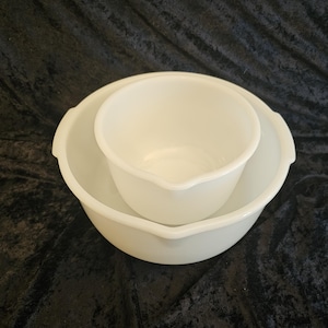 May include: Two white milk glass mixing bowls nested together. Each bowl features a smooth, opaque finish, a rounded rim, and a small integrated pouring spout on one side. The bowls have a simple, functional design with a deep basin and a wide opening.