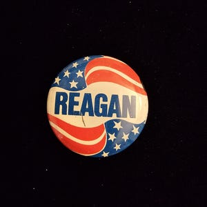K&ouml;nnte beinhalten: Ein runder Knopf mit patriotischem Design, mit roten und wei&szlig;en Streifen und wei&szlig;en Sternen auf blauem Hintergrund. Das Wort "REAGAN" ist in fetten, blauen Buchstaben in der Mitte aufgedruckt.