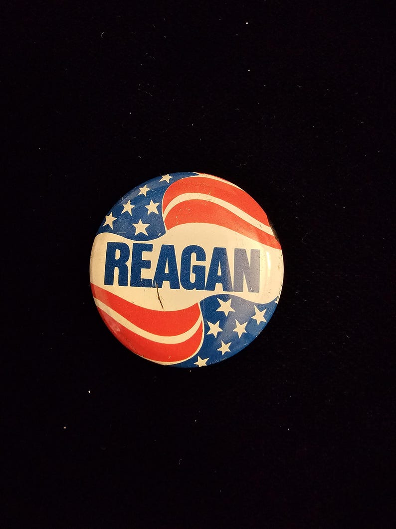 K&ouml;nnte beinhalten: Ein runder Knopf mit patriotischem Design, mit roten und wei&szlig;en Streifen und wei&szlig;en Sternen auf blauem Hintergrund. Das Wort "REAGAN" ist in fetten, blauen Buchstaben in der Mitte aufgedruckt.