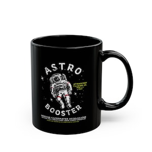 May include: Black ceramic mug featuring a white astronaut graphic and the text "ASTRO BOOSTER" in a retro style. The text "ATOMICPOP TRADING CO. NEW L.A. 2024" is also included.