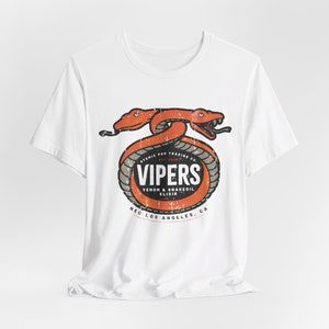 May include: White t-shirt with a graphic of two orange snakes wrapped around a circle with the text "Atomic Pop Trading Co. Est. 2024 Vipers Venom & Snakeoil Elixir Neo Los Angeles, CA"