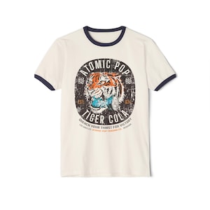 Könnte beinhalten: Ein weißes T-Shirt mit einem marineblauen Ringel. Das Shirt zeigt eine Grafik im Vintage-Stil mit einem Tiger und dem Text "Atomic Pop Tiger Cola" und "Quench Your Thirst For Victory".