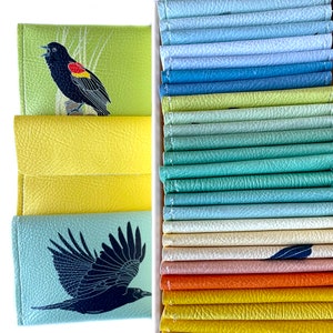 An assortment of brightly colored vegan leather wallets. Right side of the image wallets are seen from above tightly packed into a box. On the left side of the image a red-winged blackbird and crow wallet are shown