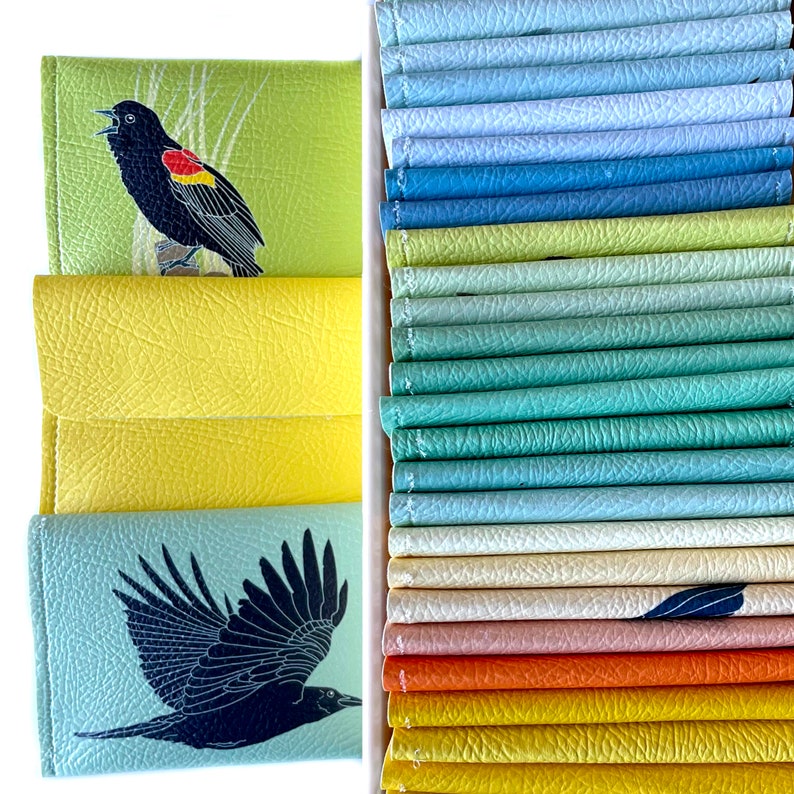 An assortment of brightly colored vegan leather wallets. Right side of the image wallets are seen from above tightly packed into a box. On the left side of the image a red-winged blackbird and crow wallet are shown