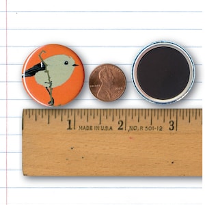 May include: A 1 inch button featuring a stylized illustration of a small bird with a black head and white body on an orange background. The button is next to a US penny and a ruler for size reference.