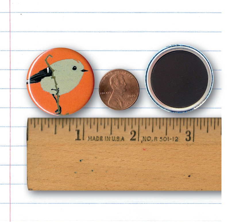May include: A 1 inch button featuring a stylized illustration of a small bird with a black head and white body on an orange background. The button is next to a US penny and a ruler for size reference.