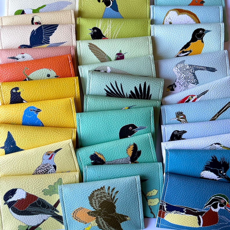 A colorful assortment of vegan leather wallets are arranged in in overlapping layers organized by colors. Each wallet is printed with a different bird illustration.