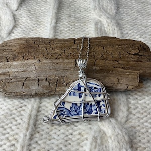 Può includere: Una collana con pendente artigianale con un pezzo di porcellana blu e bianca, avvolta in filo metallico color argento. La porcellana ha un motivo floreale. Il pendente pende da una delicata catena d'argento. La collana è esposta su legno alla deriva.