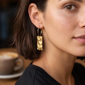 May include: A gold-toned earring featuring a thin circular hoop attached to a rectangular pendant with a textured, wavy surface. The metal has a matte finish and hangs vertically from the earlobe, reflecting light across its uneven, rippled face.