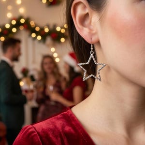 May include: A silver star-shaped earring with a textured surface hangs from a woman's ear. The earring is a festive accessory, suitable for holiday celebrations. The background shows a blurred party scene with Christmas lights.