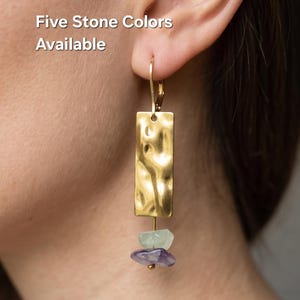 May include: A gold-toned drop earring featuring a rectangular metal plate with a hammered, textured surface. Suspended from the bottom of the plate are two small, irregular, raw-cut stones in light green and purple. Text reads: Five Stone Colors Available.