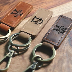 May include: Three leather keychains with metal rings and clasps. Each keychain has a personalized name and initials engraved on the leather. The keychains are brown, tan, and dark brown.