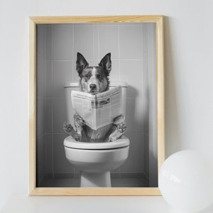 May include: A black and white photo of a dog sitting on a toilet and reading a newspaper. The dog is wearing a collar and has a serious expression on its face. The photo is framed in a light wood frame.