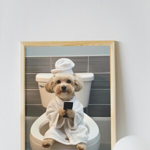 May include: A framed print featuring a small dog in a white robe and towel, sitting on a toilet while holding a smartphone. The background shows a bathroom setting with gray and blue tiles.