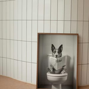 May include: A black and white photo of a dog sitting on a toilet and reading a newspaper. The photo is framed in a brown wood frame.