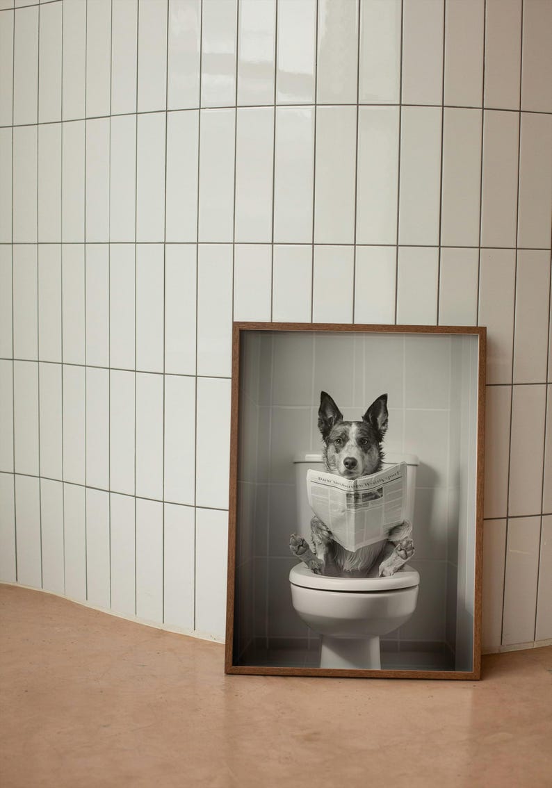 May include: A black and white photo of a dog sitting on a toilet and reading a newspaper. The photo is framed in a brown wood frame.