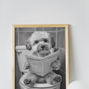 May include: A black and white photo of a dog sitting on a toilet and reading a newspaper. The dog is wearing a serious expression and is holding the newspaper with its paws. The photo is framed in a light wood frame.