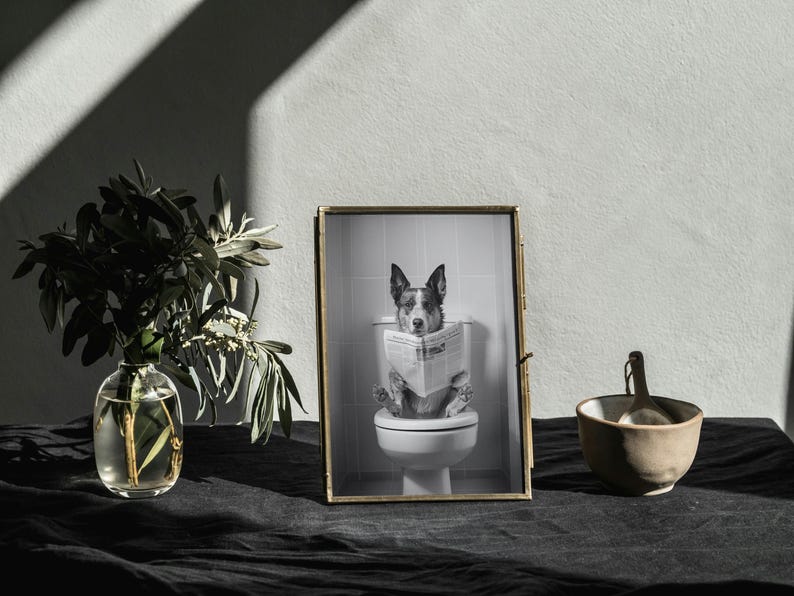 May include: A black and white photo of a dog sitting on a toilet and reading a newspaper. The photo is framed in a gold frame and is sitting on a black table.