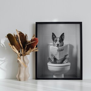 May include: Black and white photo of a dog sitting on a toilet and reading a newspaper. The photo is framed in a black frame.
