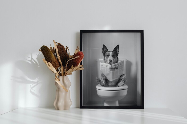 May include: Black and white photo of a dog sitting on a toilet and reading a newspaper. The photo is framed in a black frame.