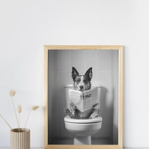 May include: Black and white photo of a dog sitting on a toilet and reading a newspaper. The photo is framed in a light wood frame.
