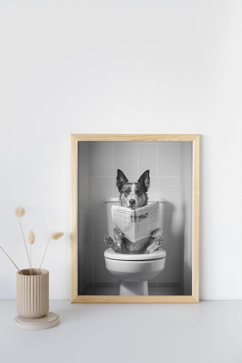 May include: Black and white photo of a dog sitting on a toilet and reading a newspaper. The photo is framed in a light wood frame.