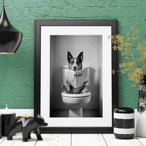 May include: A black and white photo of a dog sitting on a toilet and reading a newspaper. The photo is framed in a black frame and hanging on a green brick wall.