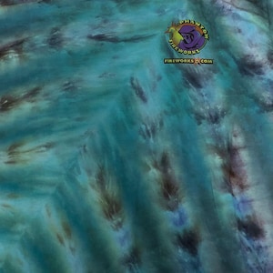 May include: A turquoise tie-dye t-shirt with a round logo on the upper left chest. The logo features a purple cartoon character and the text "Fireworks.com" in yellow and white. The shirt has a crew neck and short sleeves.