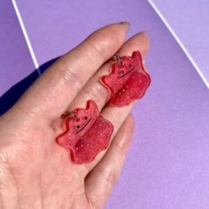 May include: Two red glittery Ditto-shaped keychains with black facial features. The keychains are made of resin and have silver-coloured metal key rings. The keychains are shown against a purple background.
