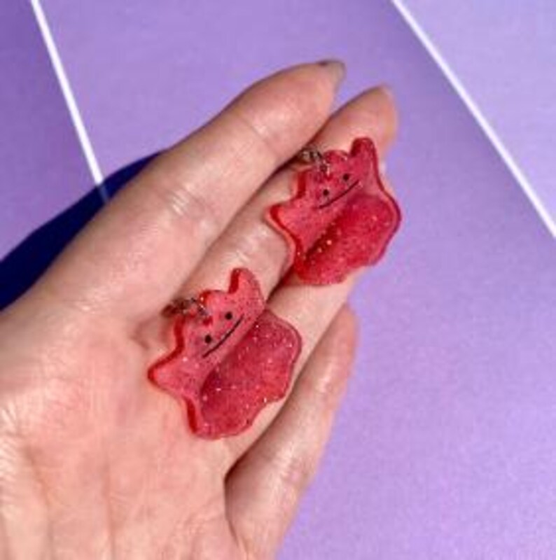 May include: Two red glittery Ditto-shaped keychains with black facial features. The keychains are made of resin and have silver-coloured metal key rings. The keychains are shown against a purple background.