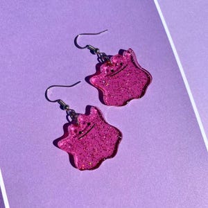 May include: Two pink glitter earrings in the shape of a cartoon character with a smiling face. The earrings have silver hooks and are set against a purple background.
