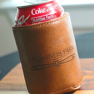 May include: A brown leather can cooler with the text "Tailwinds Farm 2023" and a propeller design. The cooler is holding a can of Coca-Cola.