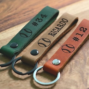 May include: Three leather keychains with baseball-themed designs. One keychain is green with the number "#34" and a baseball graphic. Another keychain is brown with the name "RICARDO" and a baseball graphic. The third keychain is orange with the number "#12" and a baseball graphic.