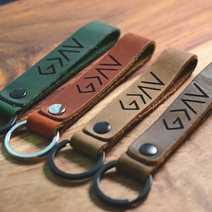 May include: Four leather keychains with black metal key rings. Each keychain has a different color: green, brown, beige, and gray. The keychains are engraved with the word "GAV" in a stylized font.