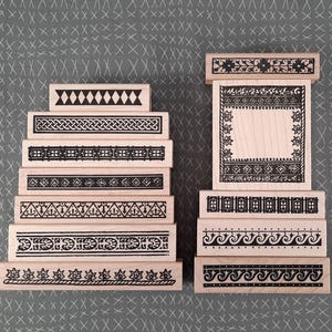 May include: A collection of wooden stamps with various black border designs. The stamps feature geometric patterns, floral motifs, and Celtic knotwork. The stamps are arranged in two stacks on a gray surface.