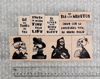 May include: Set of six wooden stamps featuring Day of the Dead imagery and text in Spanish and English. The stamps feature skeletons in various poses, including a skeleton playing a guitar, a skeleton wearing a top hat, and a skeleton wearing a traditional Japanese kimono. The text on the stamps includes phrases like "Hay mas tiempo que vida" and "He who lives with hope dies happy".