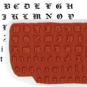 May include: A set of black gothic alphabet stamps, with both uppercase and lowercase letters. The stamps are on a red rubber base with a white border. The letters are also printed in black on the white background.