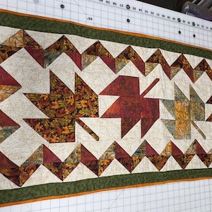 May include: A rectangular autumn-themed quilt featuring leaf patterns in shades of red, orange, and brown. The quilt has a cream-colored background with a green border and measures approximately 18 inches wide and 40 inches long.