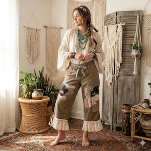 May include: A woman wearing a cream-colored blouse with embroidered details and wide sleeves, paired with olive green patchwork pants. The pants have ruffle trim at the ankles. She is accessorized with turquoise necklaces and bracelets, in a bohemian style.